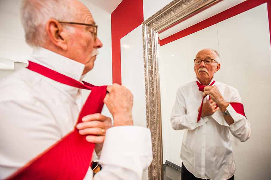 Die REHA nach der Schlaganfall-REHA - Älterer Mann bindet sich rote Kravatte vor großem Spiegel